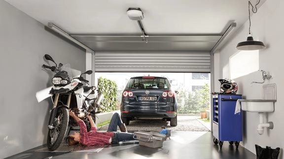 Mann repariert Motorrad in Garage, Auto im Hintergrund, Werkzeugwagen und Waschbecken