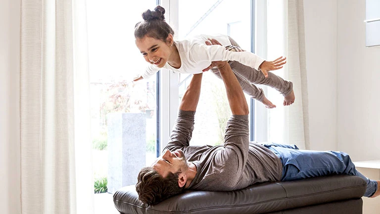 Vater spielt mit Tochter im Wohnzimmer, hebt sie lachend in die Luft