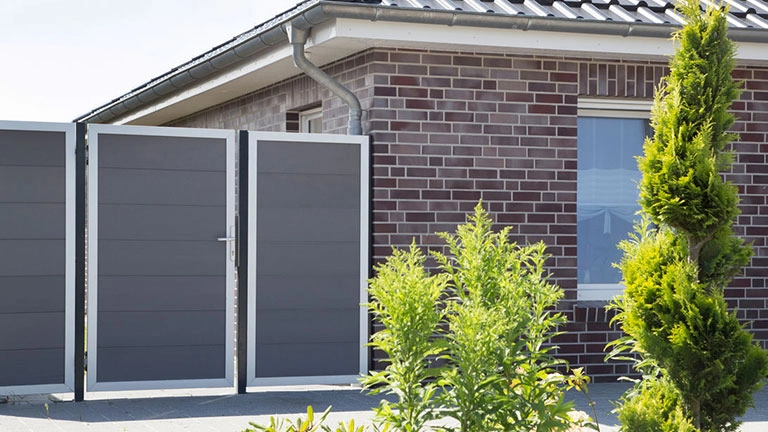 Moderner grauer Sichtschutzzaun mit Gartentor vor Backsteinhaus und Gartenpflanzen